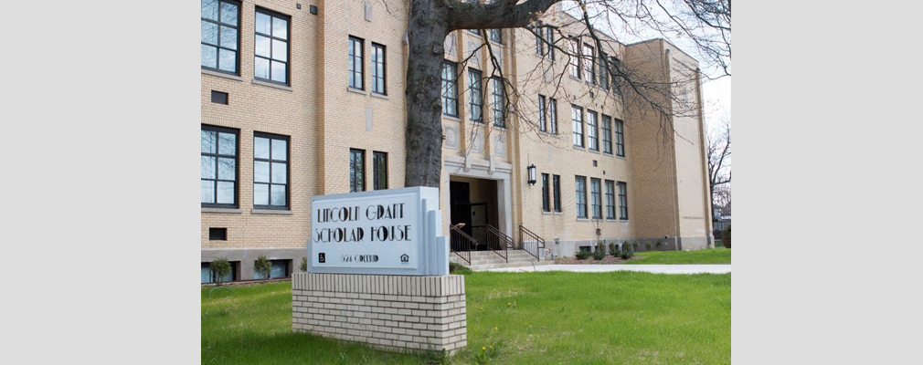 Photograph of the front façade of a three-story school building, with a freestanding sign in the foreground reading “Lincoln Grant Scholar House.”