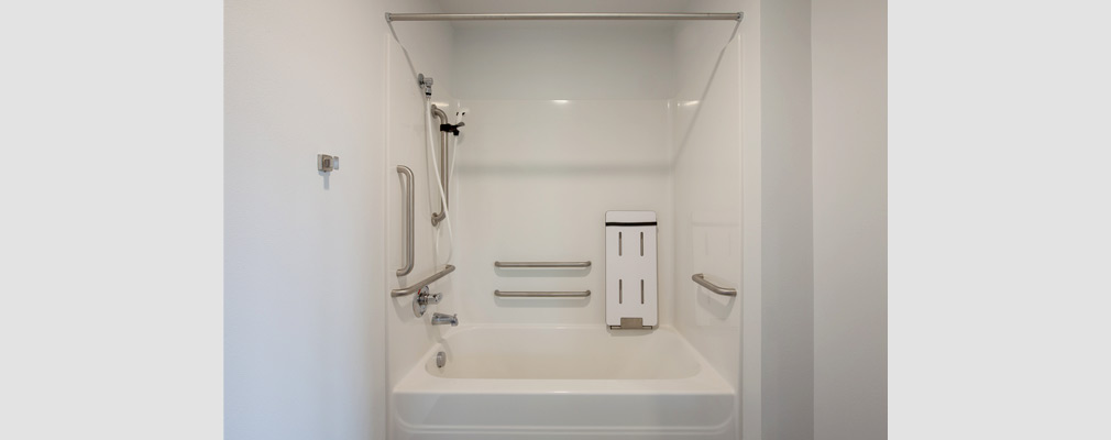 Photograph of a bathroom with a bathtub/shower combination, handrails on each wall of the tub surround, and a folding seat.