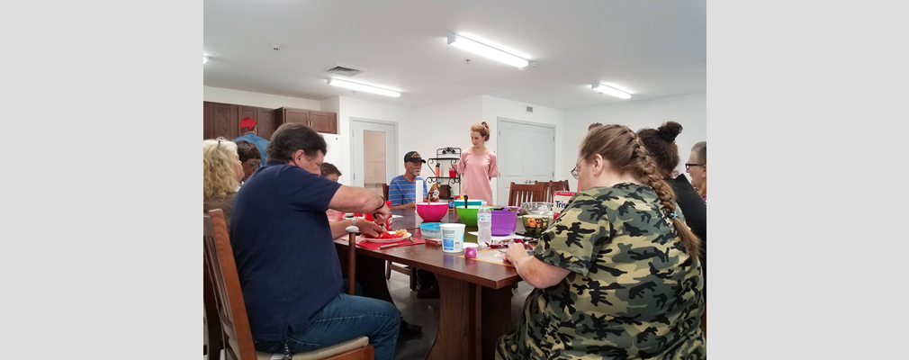 Photograph of several people in a large room preparing food around a table holding bowls and other food containers. 