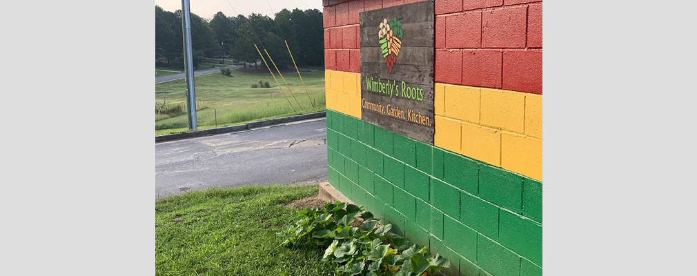 Photograph of a sign with a black background that reads “Wimberly’s Roots. Community. Garden. Kitchen.” mounted on an exterior wall of a cinder block building painted in the Pan-African colors.