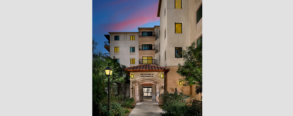 Photograph of an arched entrance to the residential component of a four-story building, with a wall sign over the entrance reading, “The Gateway at Willowbrook.” 