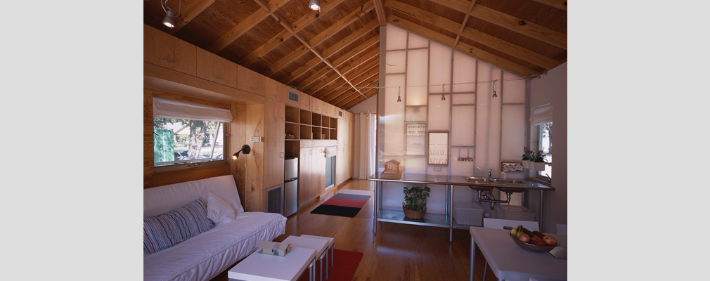 Photograph of a house interior, with a living room and kitchen in the foreground and a walkway lined with built-in shelves and cabinets in the background.