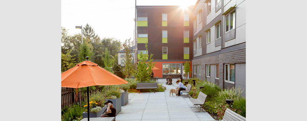 Photograph of five people and a dog in a landscaped courtyard furnished with several benches, raised planters, and tables with umbrellas.