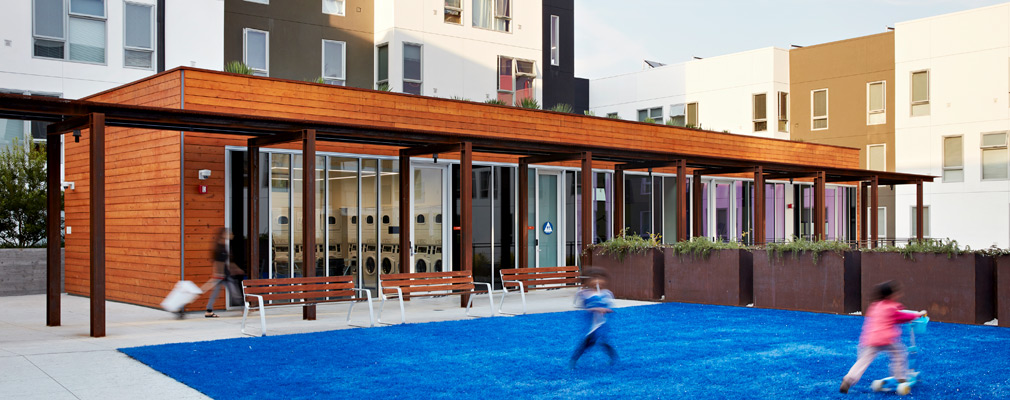 Photograph of two children playing on artificial turf in front of a one-story building with glass sliding doors; a three-story apartment building in the background frames these courtyard facilities.