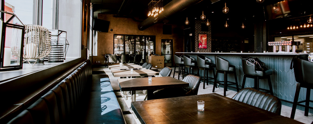 Photograph of restaurant tables with bench seating and chairs and a bar with upholstered stools.