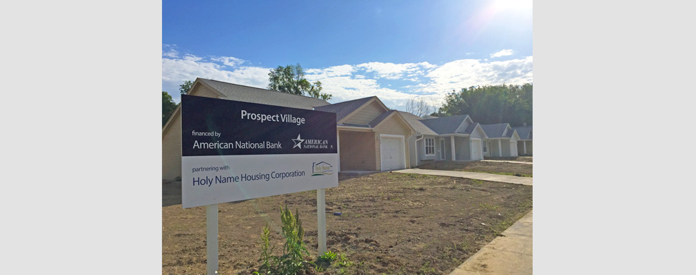 Photograph of the front façades of three newly constructed houses, with a sign in the foreground reading “Prospect Village: financed by American National Bank partnering with Holy Name Housing Corporation.”