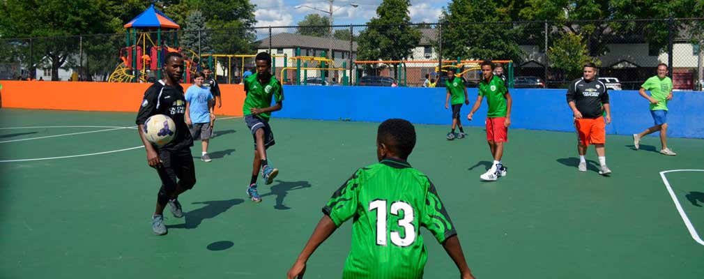 Photograph of nine adults and youth playing soccer on an asphalt court surrounded by a low wall.