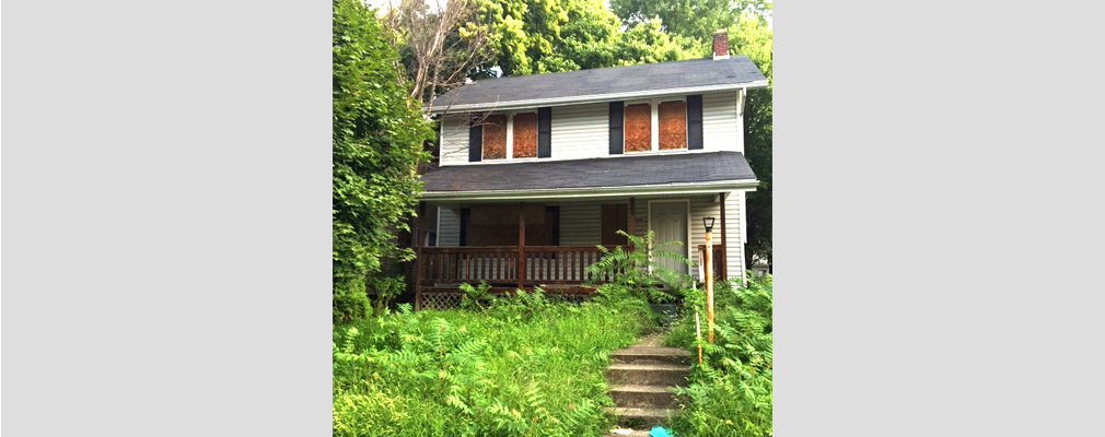 Photograph of a two-story house in poor condition, with an overgrown front yard and boarded-up windows. 