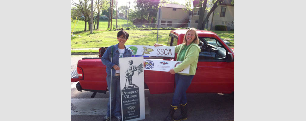 Photograph of two women in front of a pickup truck holding three organizational signs, including one that reads, “Prospect Village Neighborhood Association: Rooted with Compassion.”