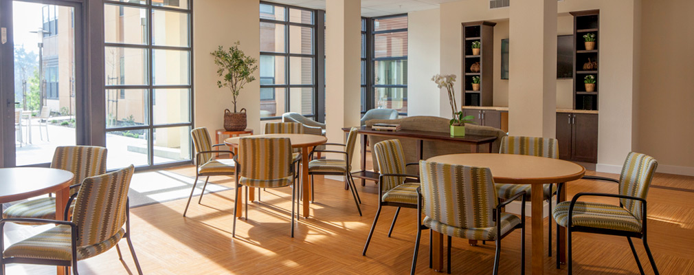 Photograph of a common area set with three circular tables with chairs, a sofa near a television, and floor-to-ceiling windows. 