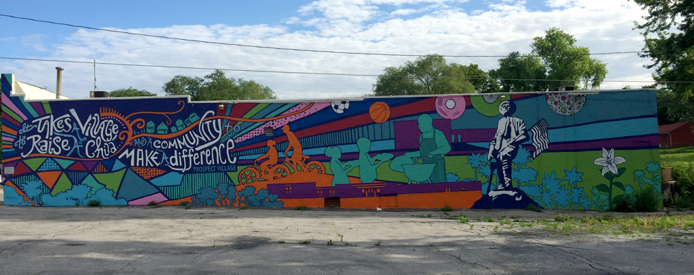 Photograph of the side façade of a one-story commercial building covered by a mural that includes “It Takes a Village to Raise a Child and a community to make a difference: Prospect Park.”