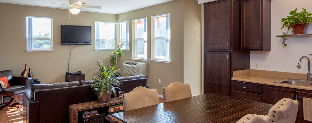 Photograph of the interior of an apartment with a dining table and chairs in the dine-in kitchen in the foreground and a sofa, chair, and windows in the living area in the background.