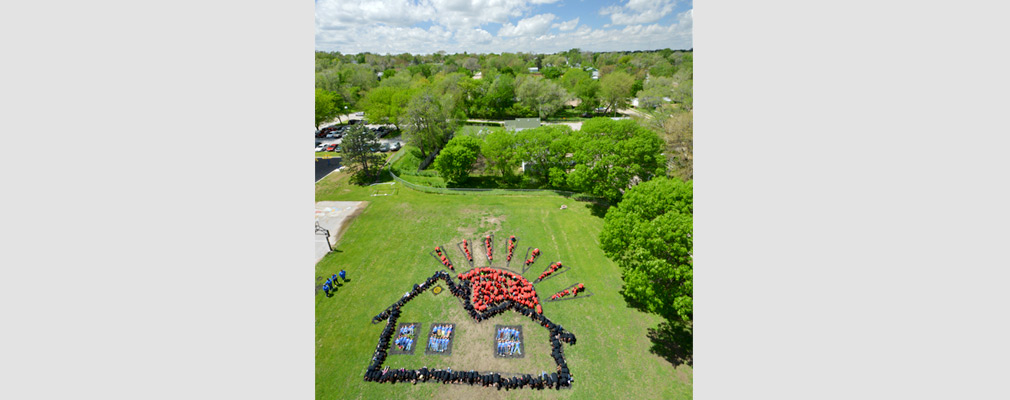 Low-altitude aerial photograph of Franklin Elementary School students in red, blue, and black shirts, lying on the ground in the shape of a house and a rising sun for the Art for the Sky project. 