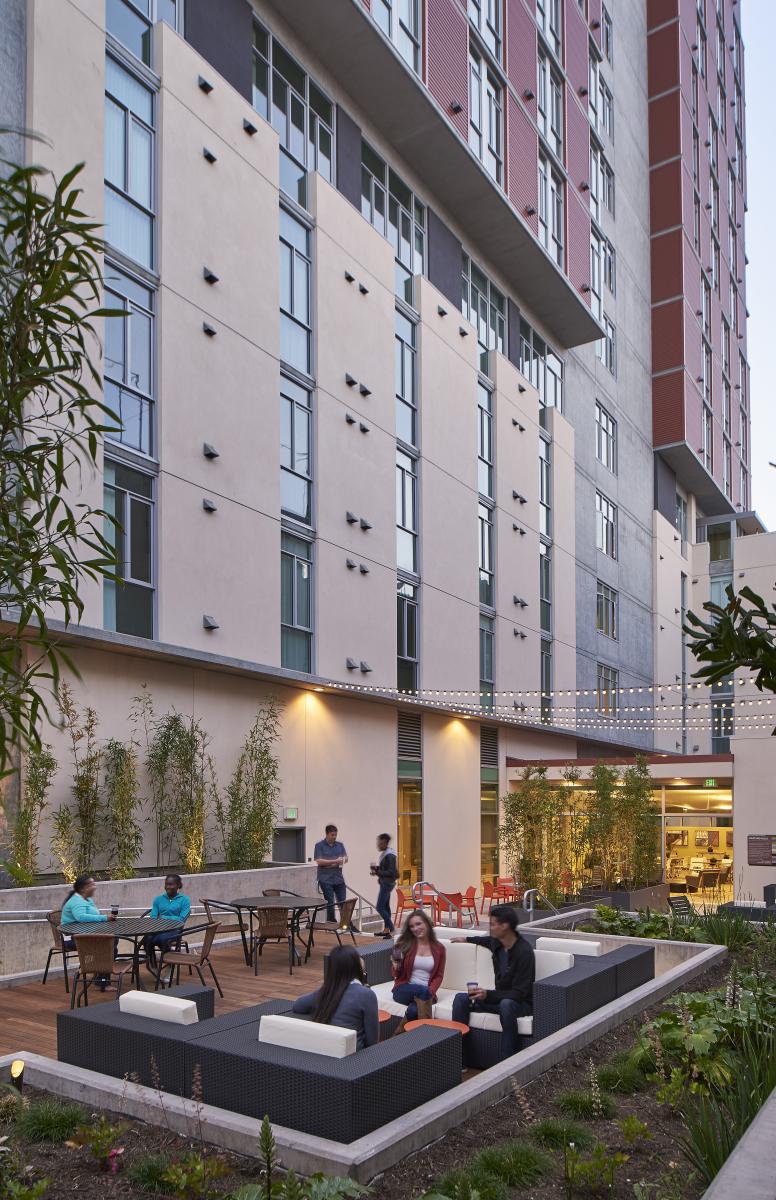 Photograph of people sitting at several tables and lounges in a paved and landscaped area next to a multifamily building.