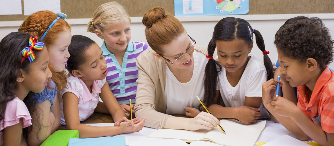 Seven young children and a teacher sit looking at a school workbook around a round table.