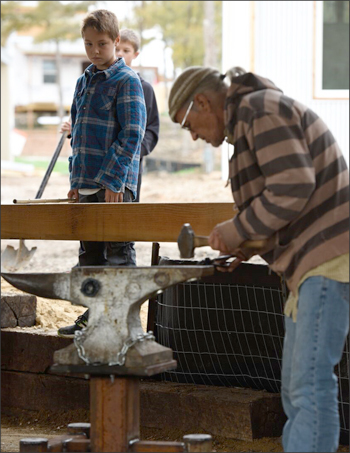 Photograph of a man working at an anvil in the community forge, with two youth watching. 