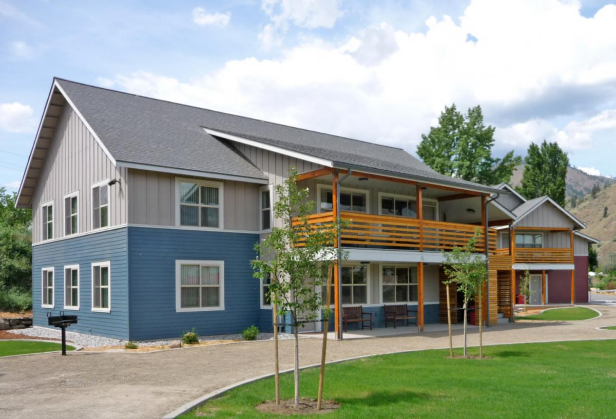 Photograph of the front and side façades of a two-story residential building featuring a patio on the first floor and a porch directly above it; another residential building is in the background.