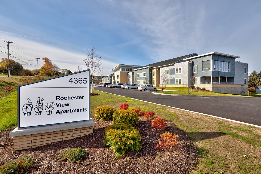 Photograph of a two-story residential building with a monument sign in the foreground reading “Rochester View Apartments” and featuring American Sign Language signs for “R,” “V,” and “A.”