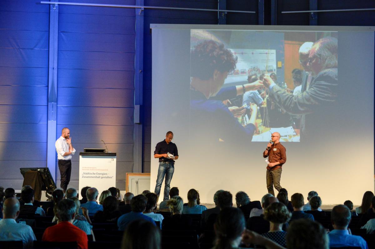 Three presenters stand onstage next to a podium in front of a pictured projected on a screen. A seated audience is visible in the foreground.