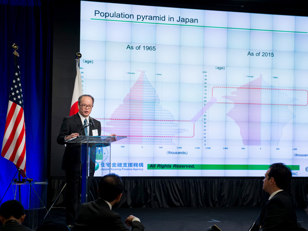 Image of a man standing behind a podium in front of a screen with two charts displaying the distribution of the population of Japan by age in 1965 and 2015.