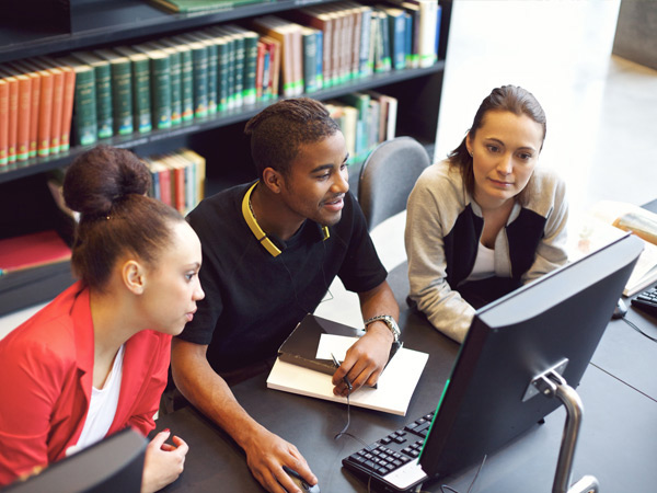 Image of three individuals sitting at a table and looking at a computer in library.