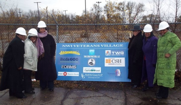 Six people in winter coats, five of them wearing hard hats, stand beside a sign listing the supporters of the NWI Veterans Village.