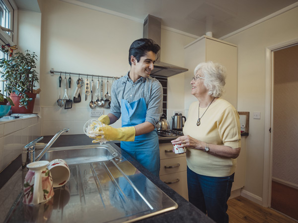 A young man cleans dishes in the kitchen and smiles at an older woman standing nearby.