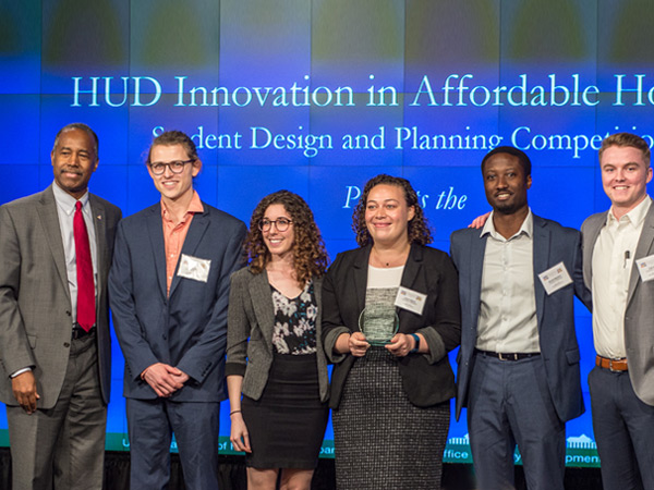HUD Secretary Ben Carson (far left) stands next to five students from the University of Maryland, College Park on stage in front of a banner stating “HUD Innovation in Affordable Housing Student Design and Planning Competition.”