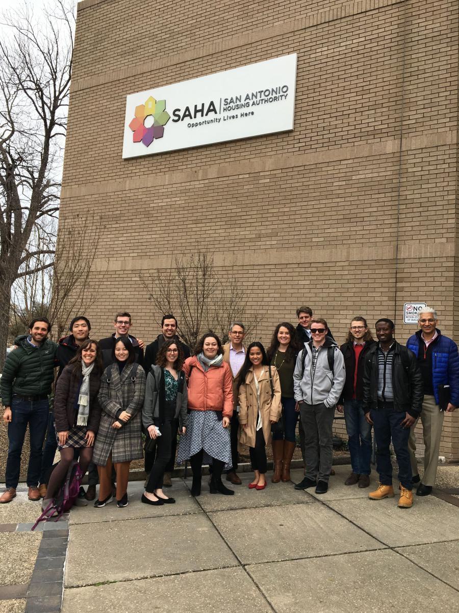 Students in the four IAH finalist teams stand in front of a building bearing a sign that says: “Opportunity Home| Opportunity Home San Antonio: Opportunity Lives Here.”