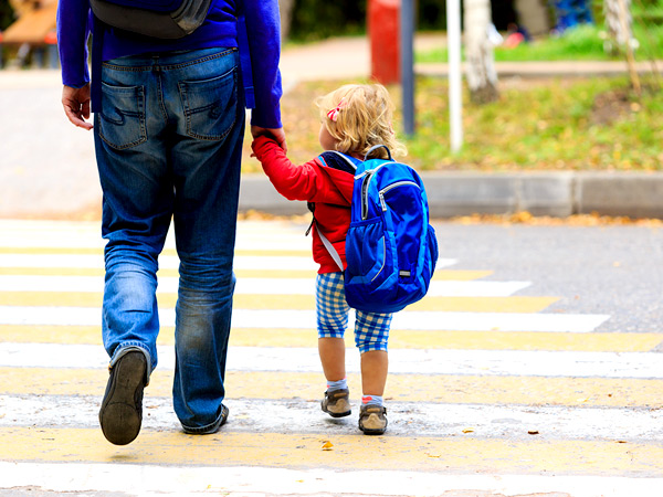 A small child with a backpack holds hands and walks with her parent outside.