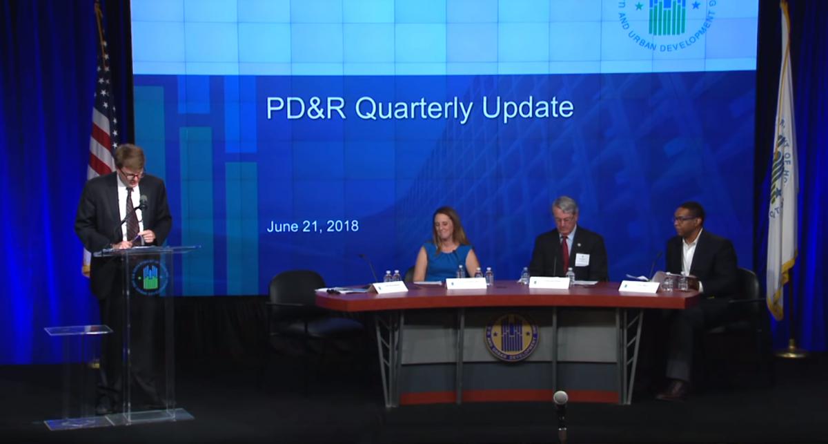 Todd Richardson, general deputy assistant secretary for PD&R, stands at a podium next to three panelists seated at a table. A screen with the words "PD&R Quarterly Update" is visible in the background. Todd Richardson, general deputy assistant secretary for PD&R, stands at a podium next to three panelists seated at a table. A screen with the words "PD&R Quarterly Update" is visible in the background.