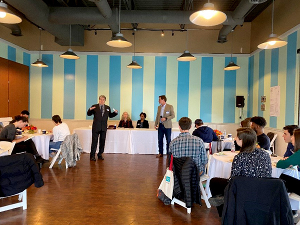 Students sit at tables and listen to a presenter standing in front of table.