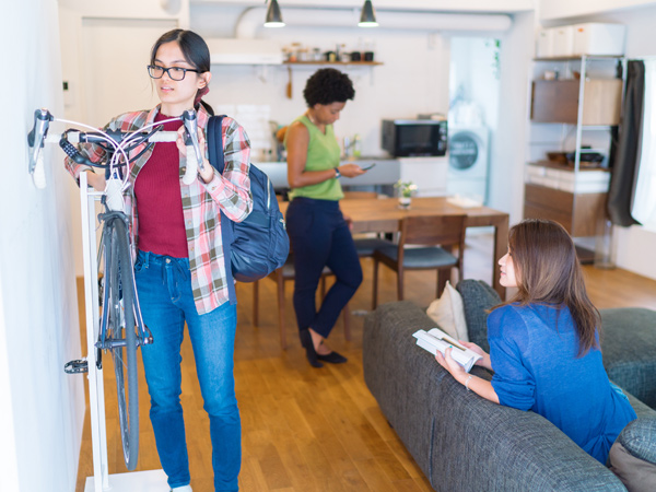 Inside an open concept room with a kitchen, dining table, and couch, one woman hangs a bicycle on a wall rack, another woman reads a book on the couch, and a third woman looks at her phone.