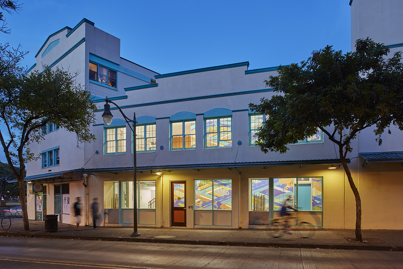 Photograph of a white stucco apartment building with pedestrians and bicyclist passing on a sidewalk. 