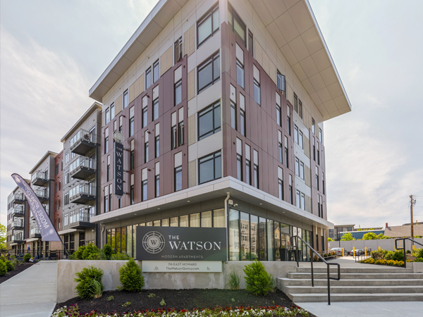 Photograph of the front and side facades of a contemporary five-story building surrounded by sidewalks and landscaping.