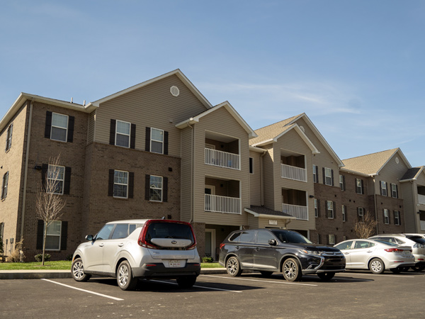 A three-story apartment building with cars and a parking lot in the forefront on a sunny day.