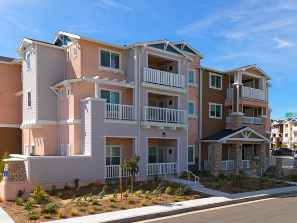 A three story apartment complex in American Farmhouse/Ranch style. Large hills and freeway signage are visible in the background.