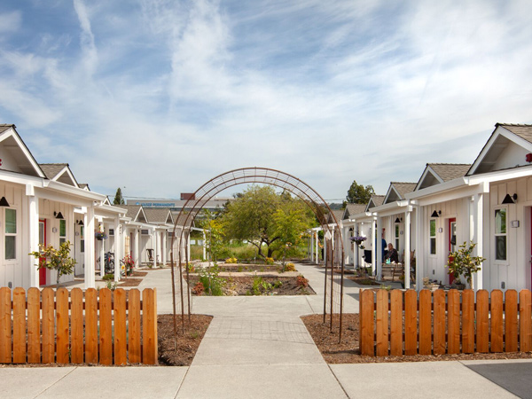 Image of an arched trellis entry way to a courtyard flanked by seven tiny homes on each side.