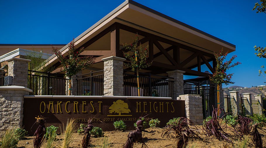 An open air entry to a building covered by a wooden patio roof in Bungalow style. In the foreground is a large wooden sign with stone pillars that reads “Oakcrest Heights” with a picture of a mature tree and address partially obscured by small plants.