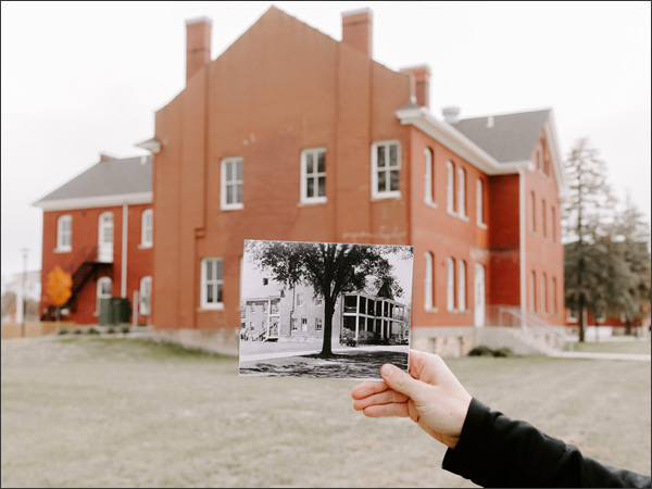 Photograph of a two-story red brick building, with a hand holding a historic black-and-white photograph of that same building in the foreground for comparison.