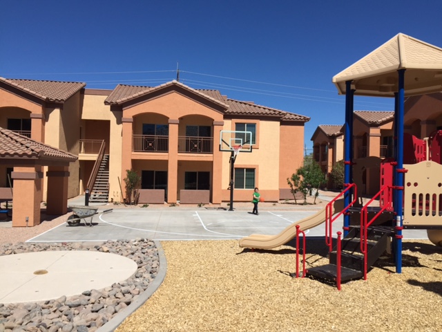 A basketball court nestled amongst two-story apartment buildings with colorful playground equipment in the foreground.