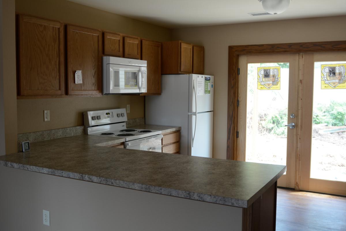 Photograph of an unfurnished kitchen area with refrigerator, oven and stovetop, and microwave.