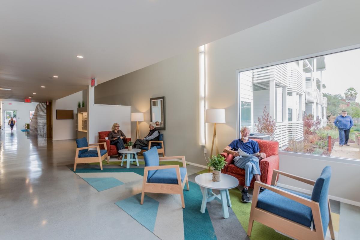 Three individuals sit in a common area with modern armchairs and tables. An individual walking along a path outside is visible through one of the windows.