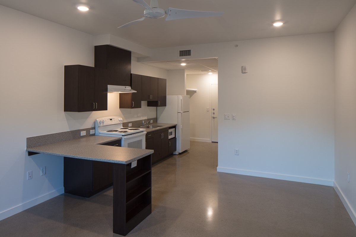 Photograph of an empty kitchen that includes countertop space, an oven, a sink, overhead cabinets, and a refrigerator.