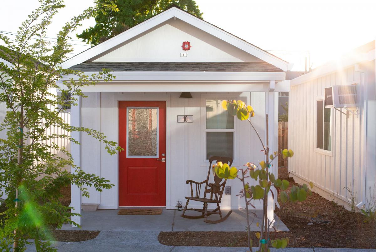 Vooraanzicht van een tiny home, wit geschilderd met een felrode deur. Een schommelstoel zit op de veranda. 