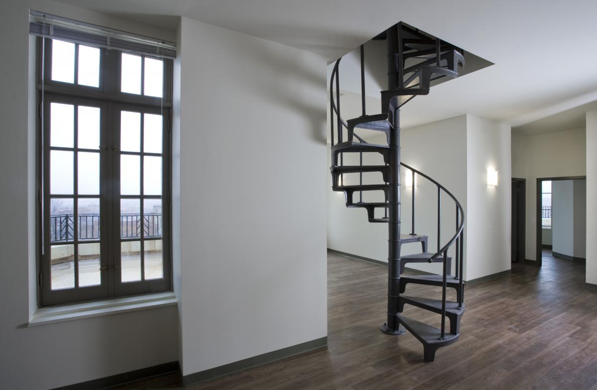 An iron spiral staircase in an unfurnished apartment with bare hardwood floors ascends up through the ceiling; an exterior window to the left looks outside.
