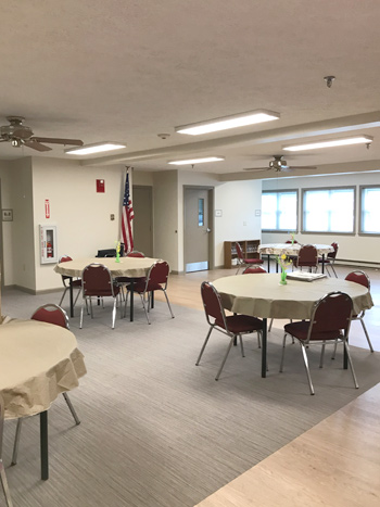 A gathering room with four tables, each with four chairs, with tablecloths and flower vases on them.