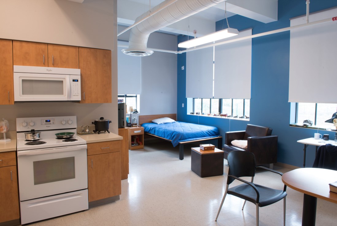 The interior of an 845 Commons residential unit. A bed and nightstand, a table and chairs, and a kitchen area with a stove, microwave, and cabinets are visible.