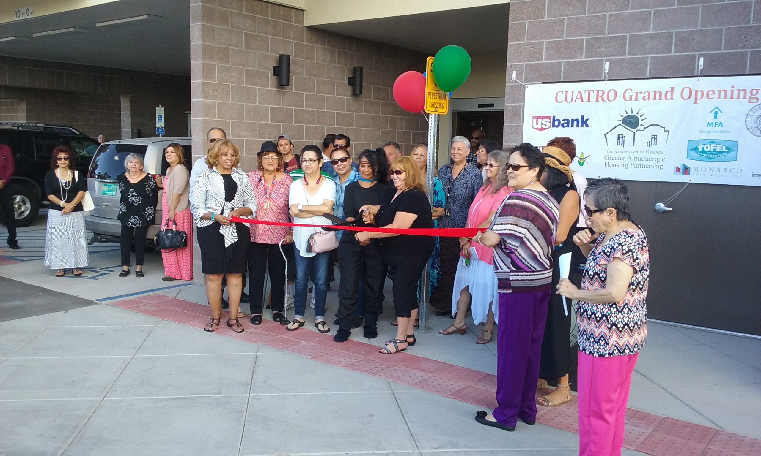 Photograph of a group of people standing in front of a building as a woman cuts a red ribbon with a large pair of scissors. 