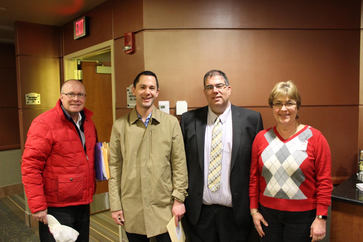 Four individuals standing in a room. A door frame is visible in the background.